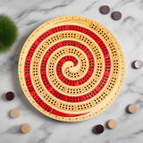 Round wooden cribbage board with red and beige spiral design on a gray background