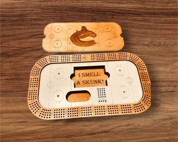 A personalized rectangular cribbage board with a hockey rink design, open to show the storage for game pieces and with a personalized engraving. A deck of cards and pegs are included.