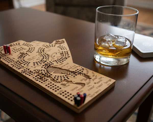 Wooden cribbage board with pegs and a glass of whiskey on a table.