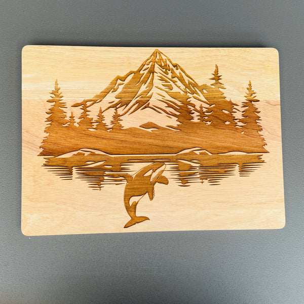 Wooden cutting board with laser-cut design of a mountain, trees, and an orca whale on a gray background