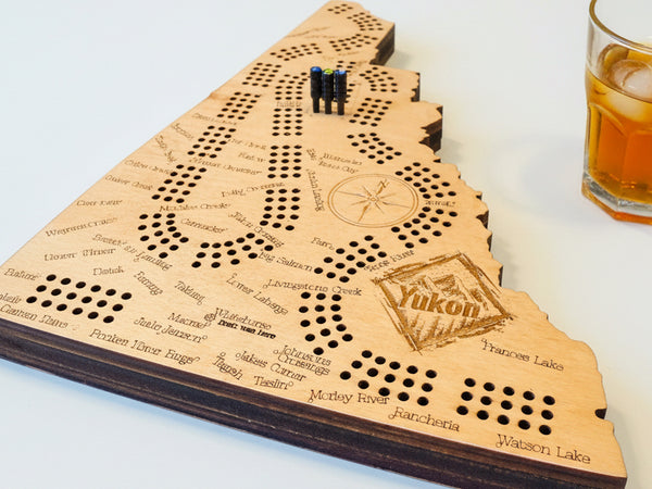 Wooden cribbage board with 'Yukon' branding on a table with playing cards and snacks.