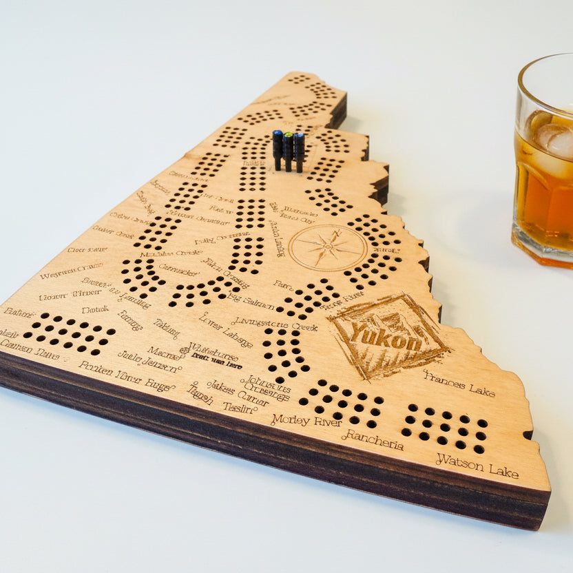 Wooden cribbage board with 'Yukon' branding on a table with playing cards and snacks.