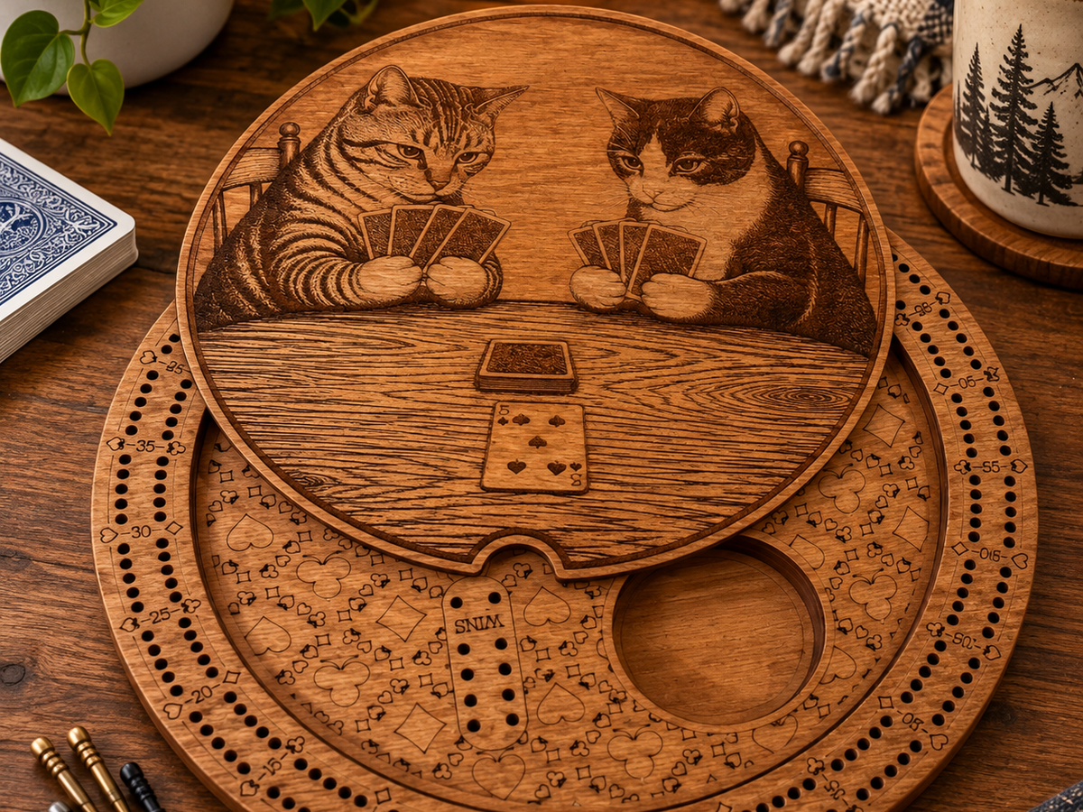 A round engraved wooden board game featuring two cats playing with cards, displayed on a wooden table with a mug, plant, and other cozy props in the background.