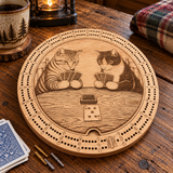 A round wooden cribbage board featuring an engraved illustration of two cats playing card games, shown on a rustic wooden table with cards, a mug, and a plaid blanket nearby.
