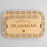 A personalized wooden cribbage board engraved with 'CHÂTEAU OKANAGAN' and a fleur-de-lis, shown on a light gray concrete surface.