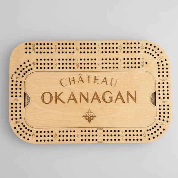 A personalized wooden cribbage board engraved with 'CHÂTEAU OKANAGAN' and a fleur-de-lis, shown on a light gray concrete surface.
