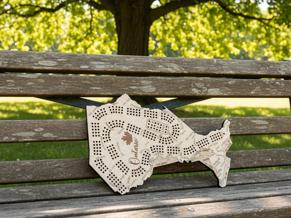 A wooden Ontario-shaped board game set displayed on a dark background, with playing cards arranged along the bottom wooden surface. The board features engraved street names, playing card suits, and the word 'Ontario' with a maple leaf.
