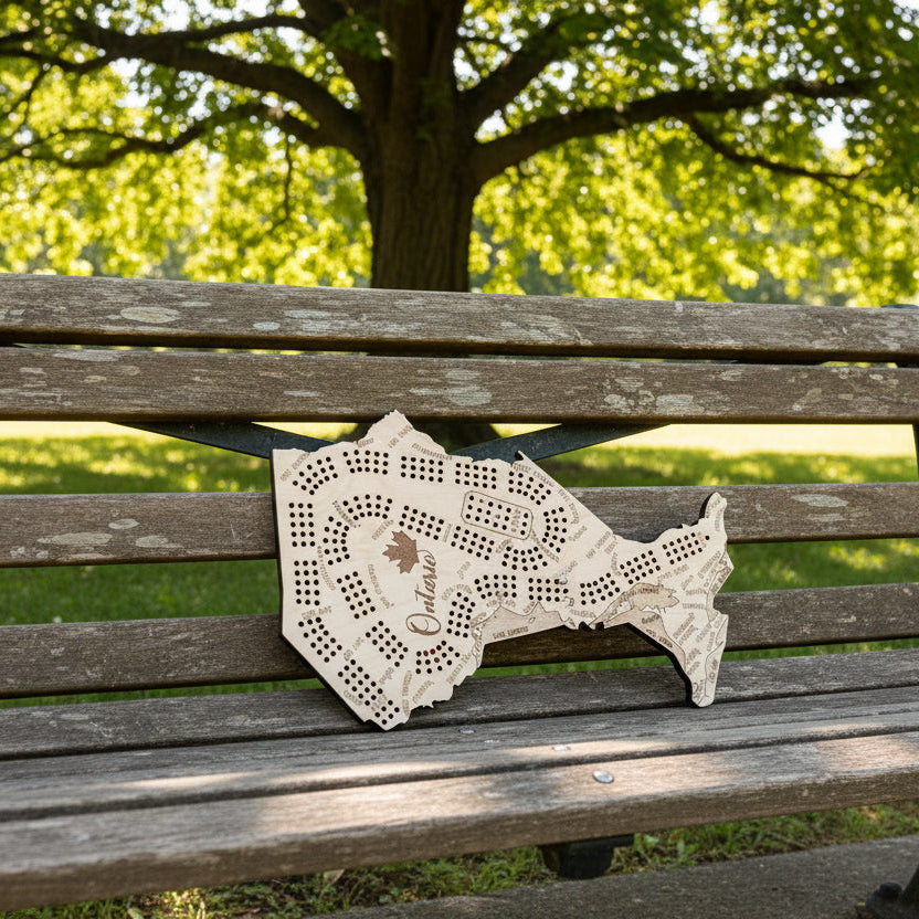 A wooden Ontario-shaped board game set displayed on a dark background, with playing cards arranged along the bottom wooden surface. The board features engraved street names, playing card suits, and the word 'Ontario' with a maple leaf.