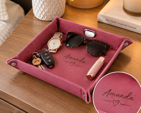 A pink personalized fabric tray with silver snap corners, placed on a wooden nightstand. The tray is decorated with the name 'Amanda' and a heart, and is shown holding various everyday items like sunglasses, a watch, keys, and a lipstick.