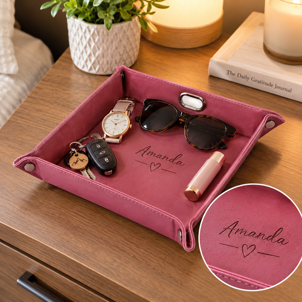 A pink personalized fabric tray with silver snap corners, placed on a wooden nightstand. The tray is decorated with the name 'Amanda' and a heart, and is shown holding various everyday items like sunglasses, a watch, keys, and a lipstick.