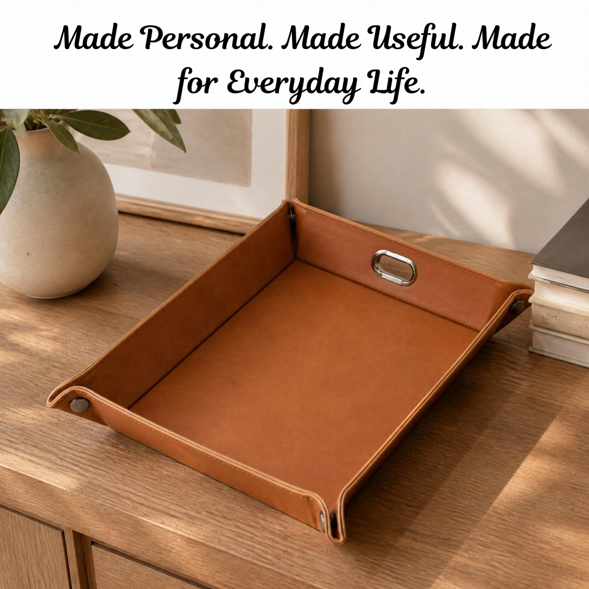 A brown leather tray with metal handles sits on a wooden table in a styled home setting, with a white banner at the top reading 'Made Personal. Made Useful.
